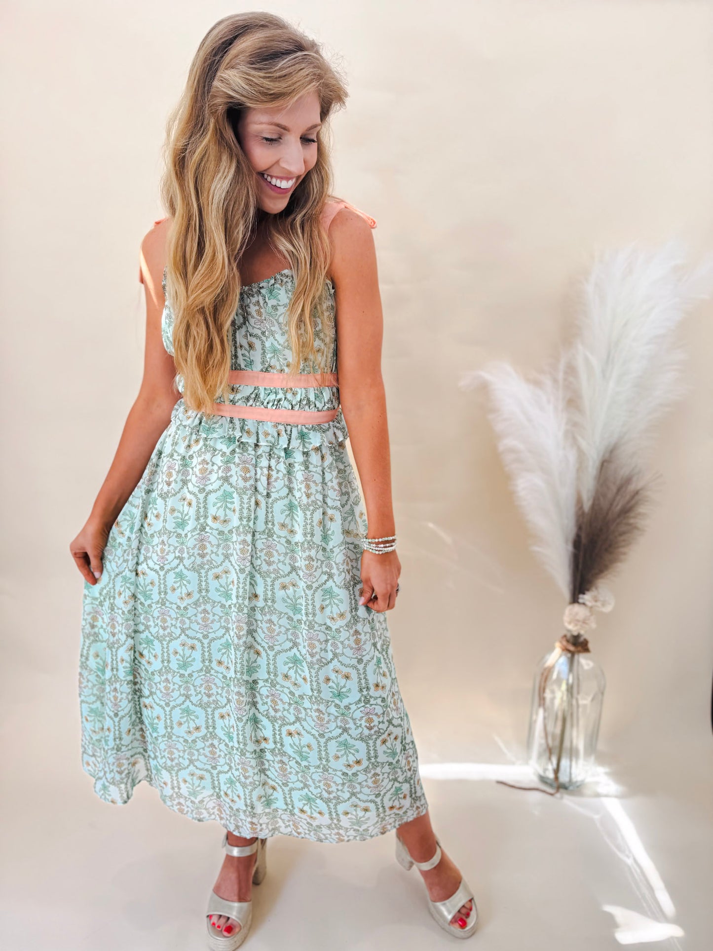 Sage green midi dress with coral ribbon worn with gold heels by model in front of a tan background