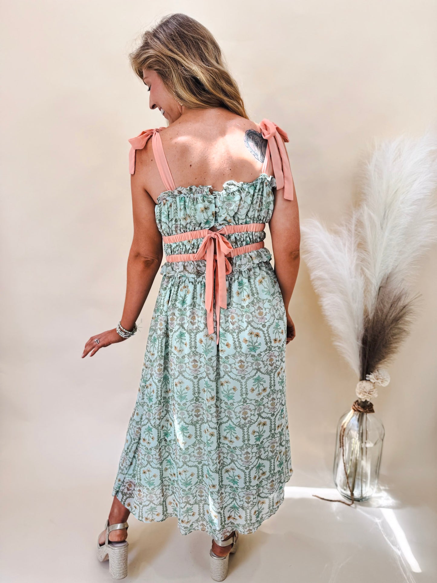 Back view of sage green midi dress with coral ribbon accents worn by model in gold heels