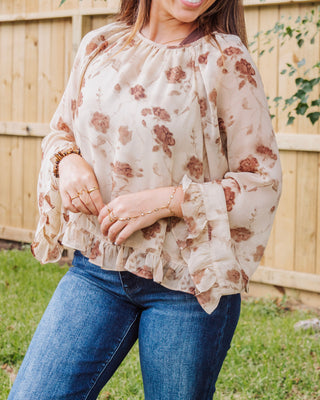 Close-up of woman wearing a beige floral chiffon blouse with ruffle hem and flared sleeves, paired with blue jeans for a soft, feminine boho look.