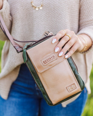 Close-up of a woman holding a Bedstu leather crossbody bag in tan and green with front pocket and stitched detailing, styled with casual jeans and sweater.