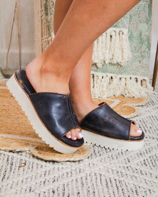 Close-up of black Bed|Stu leather platform slides with white lug sole, styled on foot in a boutique setting with woven and floral textures.