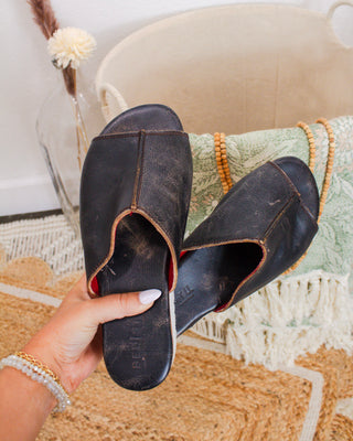 Close-up of black Bed|Stu leather slide sandals with natural wear, held by hand in boutique setting with woven decor and soft green textiles.