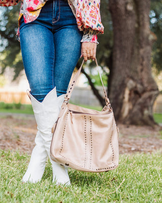 Woman wearing colorful blouse, blue jeans, and white cowgirl boots holding a tan Bed|Stu leather studded shoulder bag outdoors.