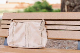 Beige Bed Stu leather crossbody bag with brass stud detailing, displayed on a wooden bench. Stylish handcrafted bag for everyday wear or boho-chic outfits.
