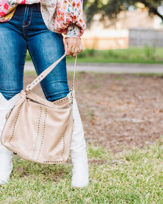 Woman holding a beige genuine leather studded shoulder bag paired with blue skinny jeans, floral blouse, and white tall boots for a chic boho look.