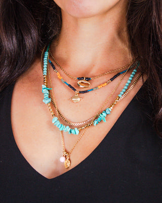 Close-up of a layered necklace stack featuring turquoise beads, gold chains, colorful seed beads, and unique celestial pendants styled with a black V-neck top.