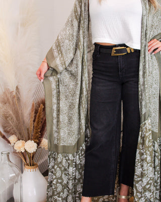 Close-up of woman wearing an olive green paisley kimono over a white sleeveless top and black wide-leg jeans, styled with a statement belt and pampas grass decor.
