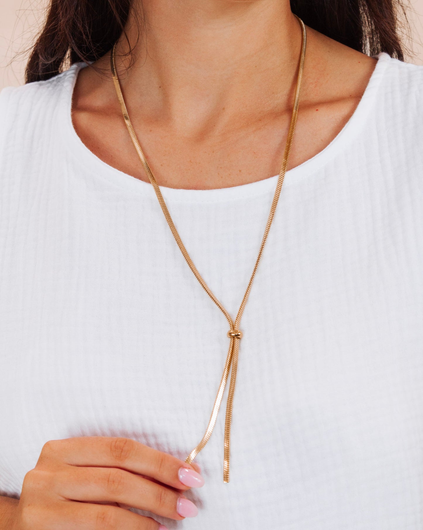 Woman wearing a gold lariat necklace with a knotted drop detail, paired with a white textured top and pink manicure, showcasing a simple and elegant jewelry look.