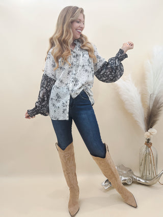 Woman wearing a mixed-print long sleeve blouse with lace detail, dark wash skinny jeans, and tan embroidered cowboy boots, posing in a stylish Western outfit.