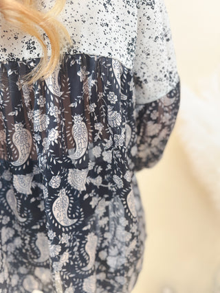 Close-up of a mixed-print blouse featuring a contrast of white and black speckled fabric with sheer black paisley floral print, showcasing texture and pattern detail.