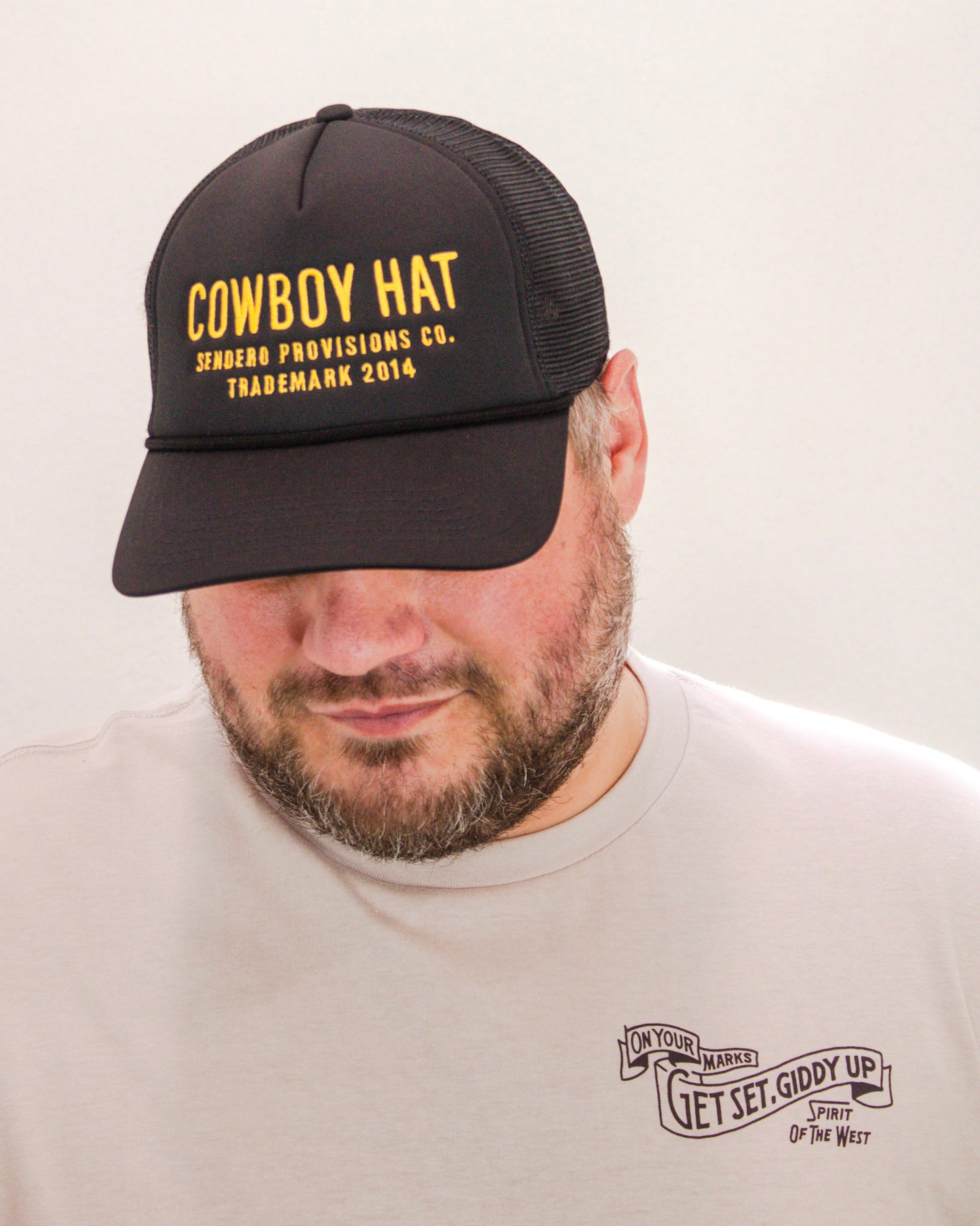 Man wearing a black mesh trucker hat with yellow “Cowboy Hat – Sendero Provisions Co.” embroidery, paired with a beige tee featuring “Get Set, Giddy Up” graphic.