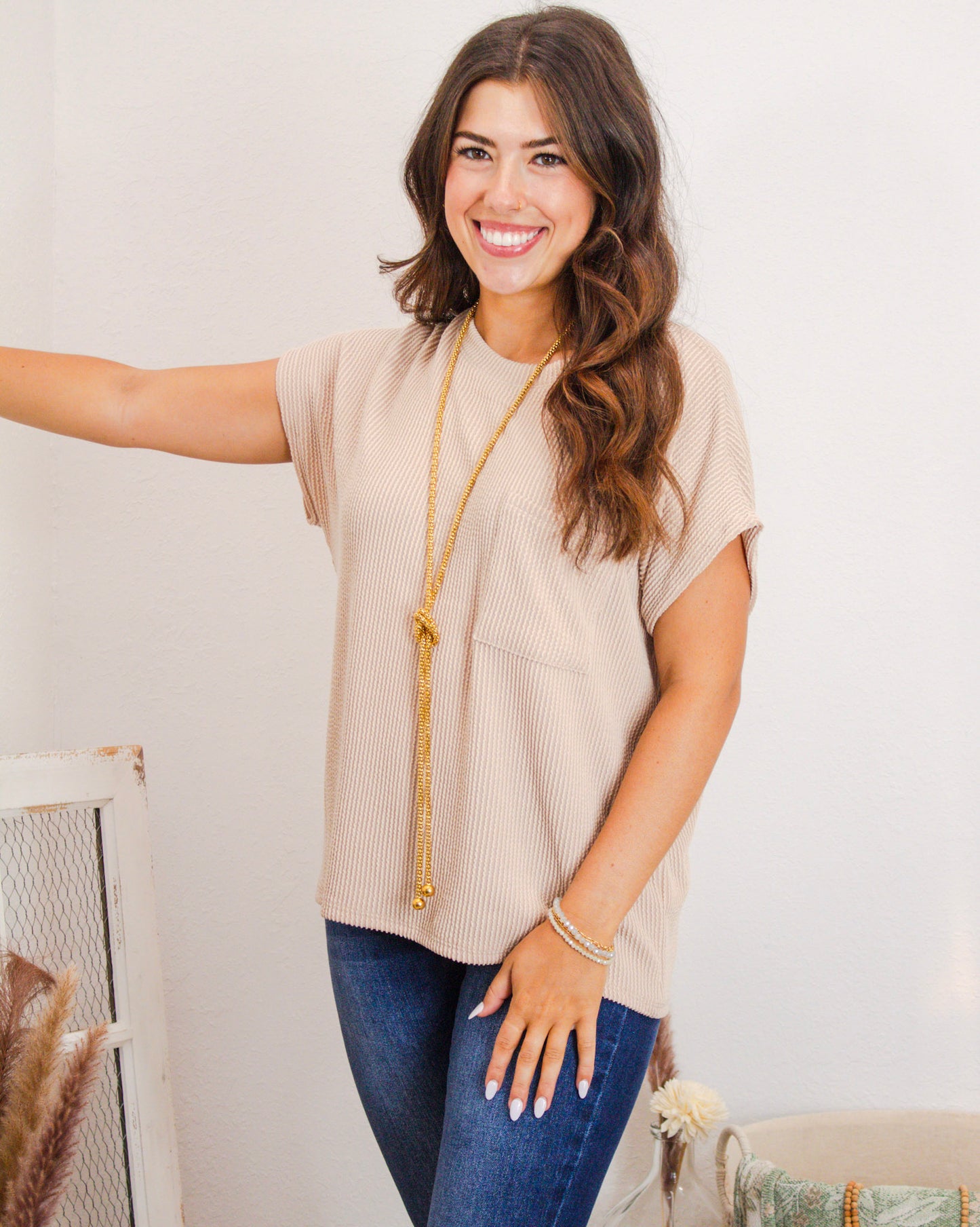 Woman wearing a beige textured short sleeve top with front pocket, styled with a long gold necklace and dark denim jeans from MaKenzie Layne Boutique.