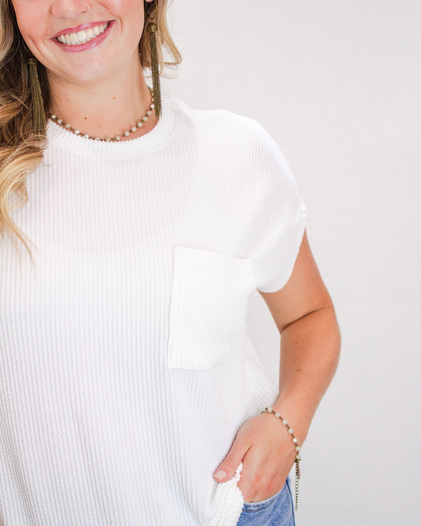 Close-up of woman wearing a textured white ribbed short sleeve top with a front pocket, paired with light wash denim jeans and beaded accessories.