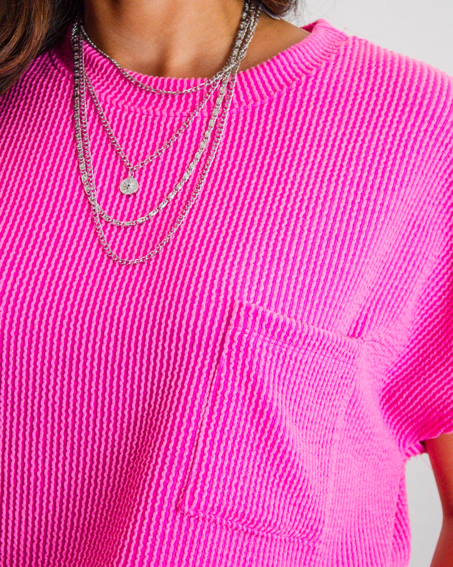 Close-up of a vibrant hot pink textured top with a front pocket, styled with layered silver chain necklaces from MaKenzie Layne Boutique.