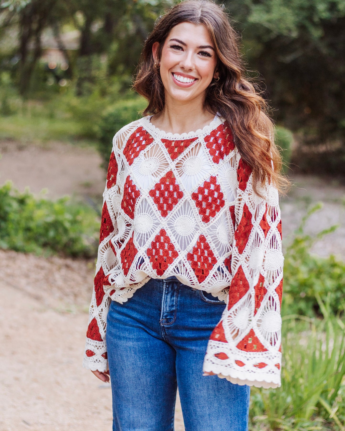 Woman wearing a long-sleeve crochet sweater with red and cream diamond patterns, paired with blue jeans, smiling outdoors in a natural setting.