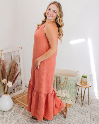 Woman modeling a flowy rust orange maxi dress with tie-strap detail and ruffle hem, styled in a cozy boho setting with soft neutral home décor.