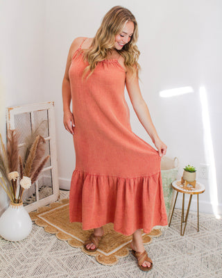 Smiling woman modeling a rust orange maxi dress with spaghetti straps and ruffle hem detail, styled in a cozy boutique corner with neutral decor.