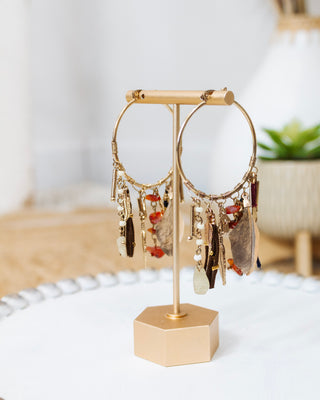 Gold hoop earrings displayed on a stand, embellished with assorted boho-style charms including feathers, beads, stones, and metallic accents.