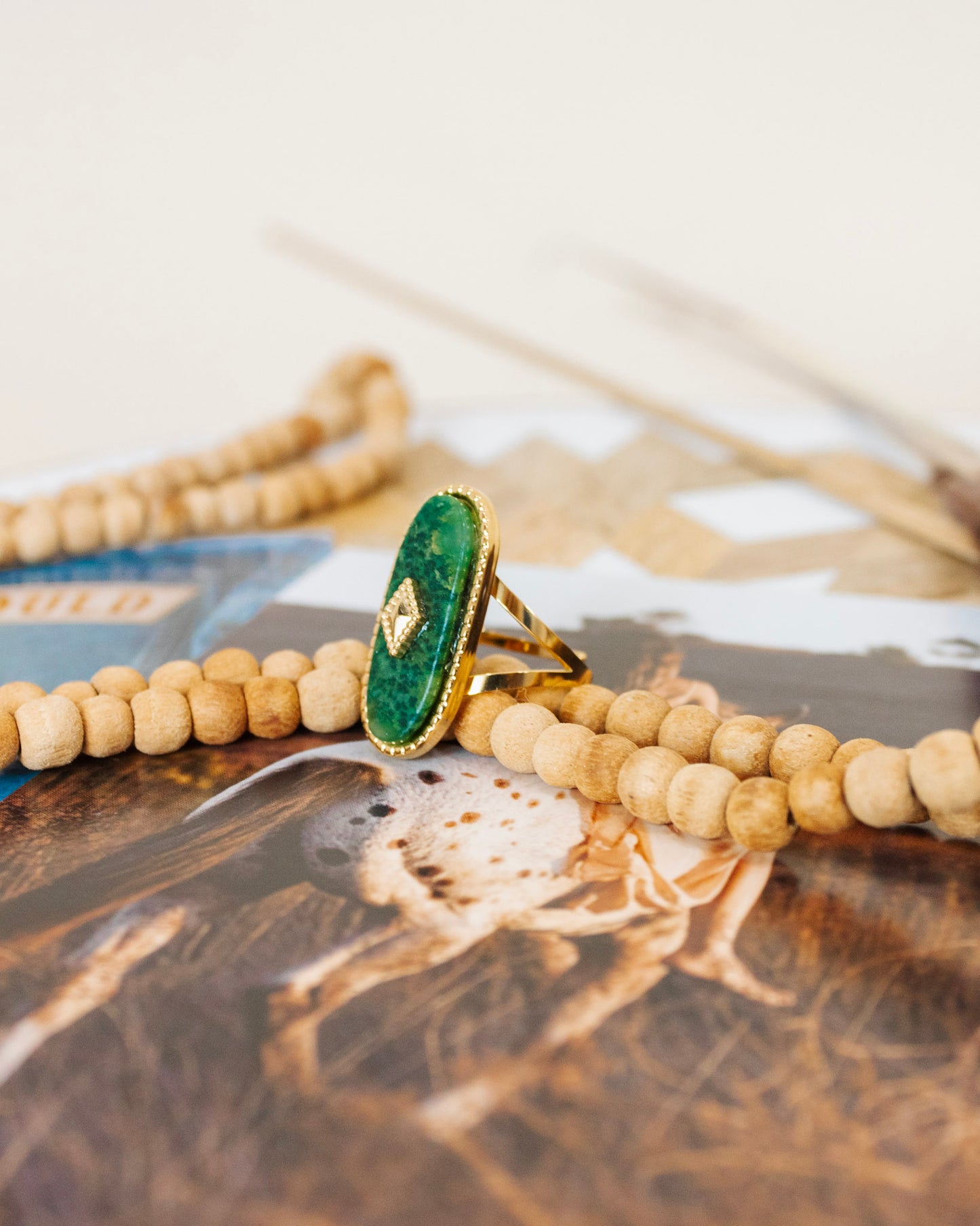 Gold ring featuring an oval emerald-green gemstone with a diamond-accented center, displayed atop natural wooden bead strands on a neutral background