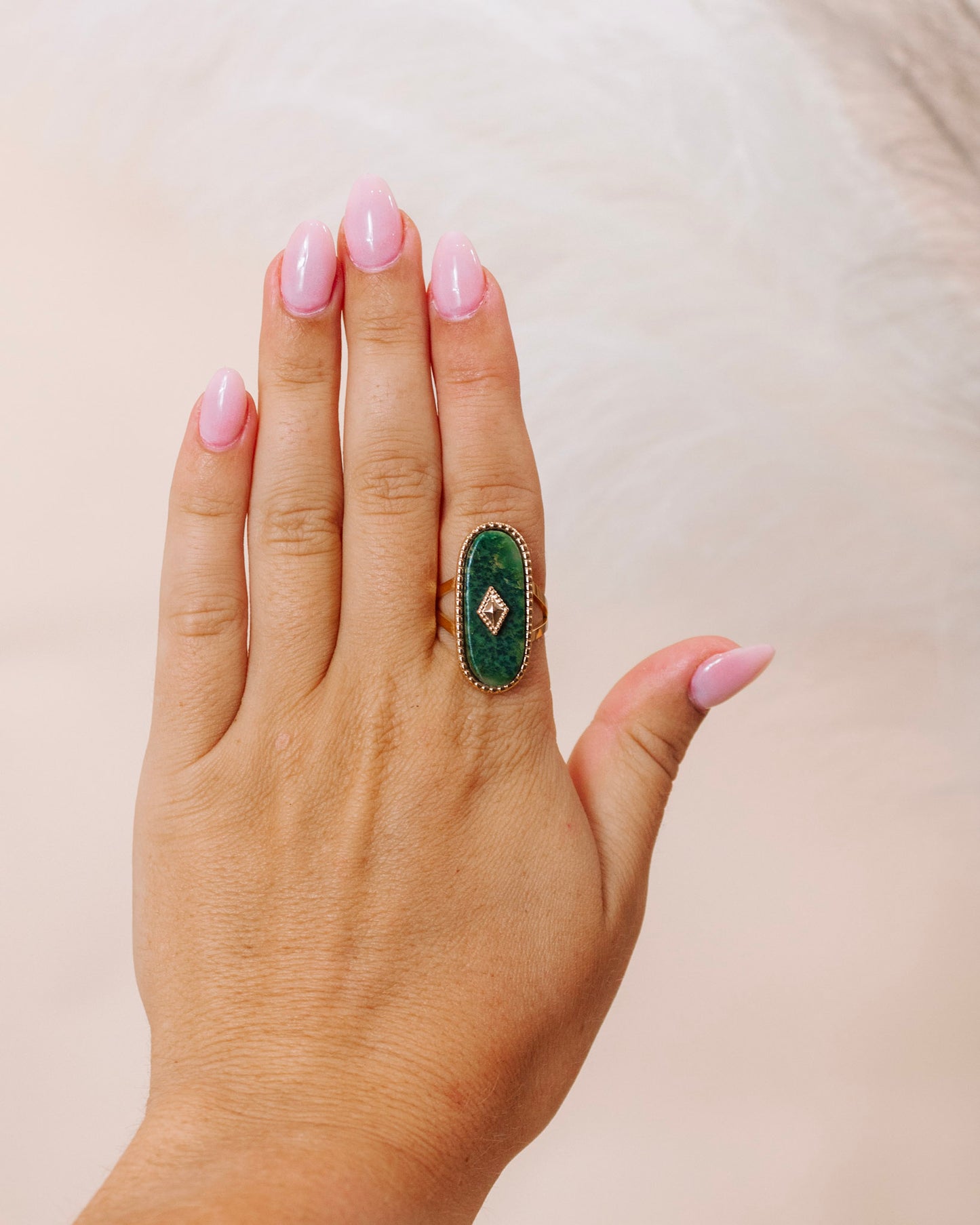 Hand wearing a gold ring with an oval green gemstone surrounded by a delicate diamond-accented bezel, styled against a soft neutral background