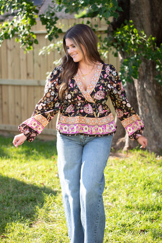 Woman wearing a boho floral wrap blouse with long sleeves in rich black, orange, and purple tones, paired with high-waisted light-wash jeans for a fall-ready look.