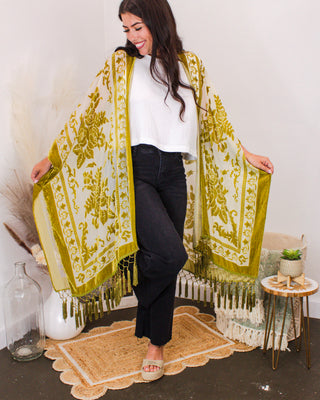 Woman wearing an ivory and olive green floral kimono with tassel fringe detail, styled with a white top and black jeans in a boho-chic room with pampas accents.