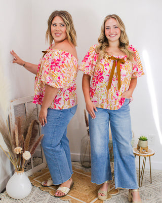 Two women wearing matching off-the-shoulder floral tops with brown bow details, paired with wide-leg denim jeans, standing in a styled boutique setting.