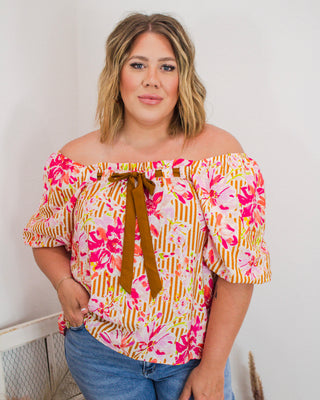 Woman wearing a colorful off-the-shoulder floral blouse with puff sleeves and a front brown tie detail, paired with light wash jeans.
