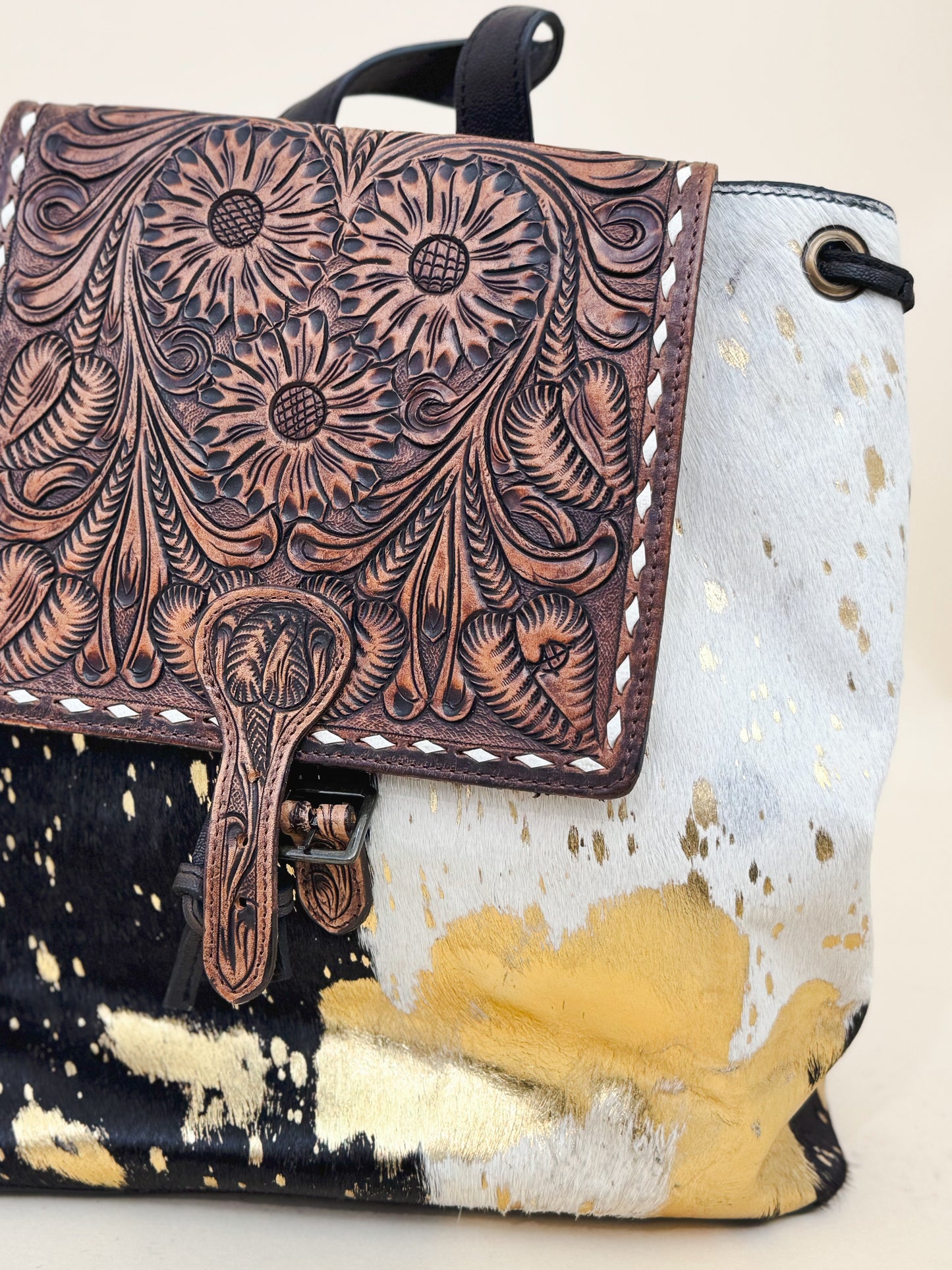 Western cowhide backpack with tooled leather floral flap and gold splatter detailing—boho-chic and rustic accessory perfect for everyday or festival style.