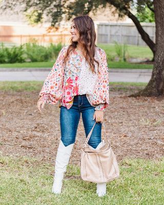Woman wearing a colorful floral patchwork boho blouse with balloon sleeves, styled with dark skinny jeans, white knee-high boots, and a beige studded tote bag.
