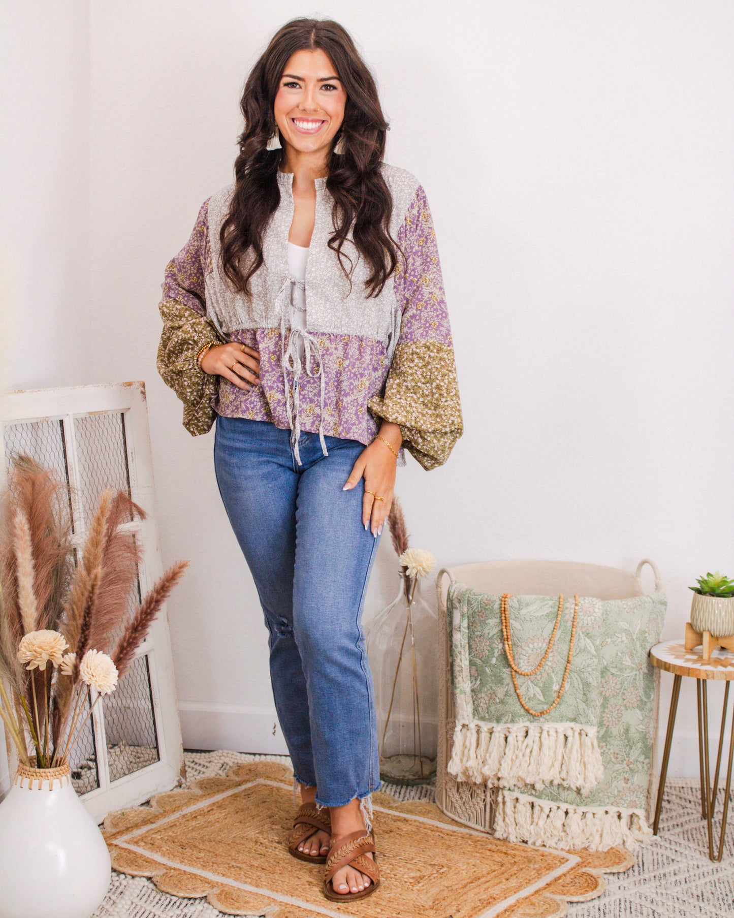 Woman wearing a colorblock boho blouse with floral sleeves and drawstring waist, paired with raw hem skinny jeans and sandals in a boutique-style room.