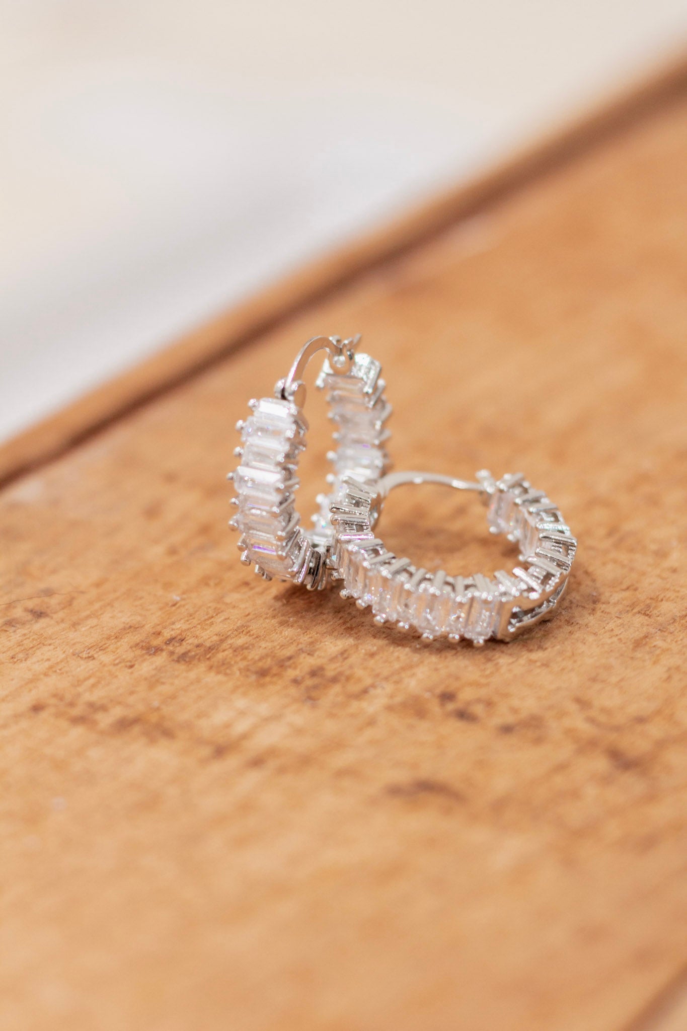 Pair of silver hoop earrings featuring baguette-cut clear crystals set in a full pavé design, displayed on a wooden surface. Glamorous and eye-catching earrings perfect for formal occasions or evening wear.