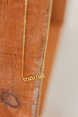 Gold necklace with lowercase 'mama' word pendant on a delicate chain, displayed against a wooden background. Minimal and meaningful jewelry gift for moms, perfect for Mother's Day or everyday wear.