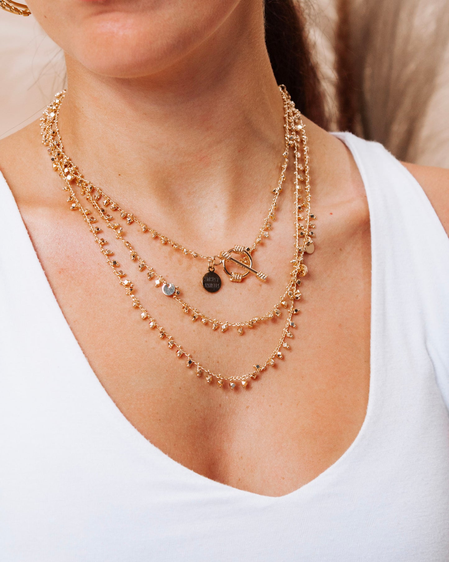 Close-up of layered gold necklaces featuring a toggle clasp, small disc charms, and dangling bead accents, styled over a white tank top