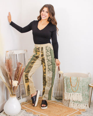 Woman modeling sage green floral & bandana patchwork jeans in a cozy boho room, wearing black V-neck top—product photo for Makenzie Layne Boutique