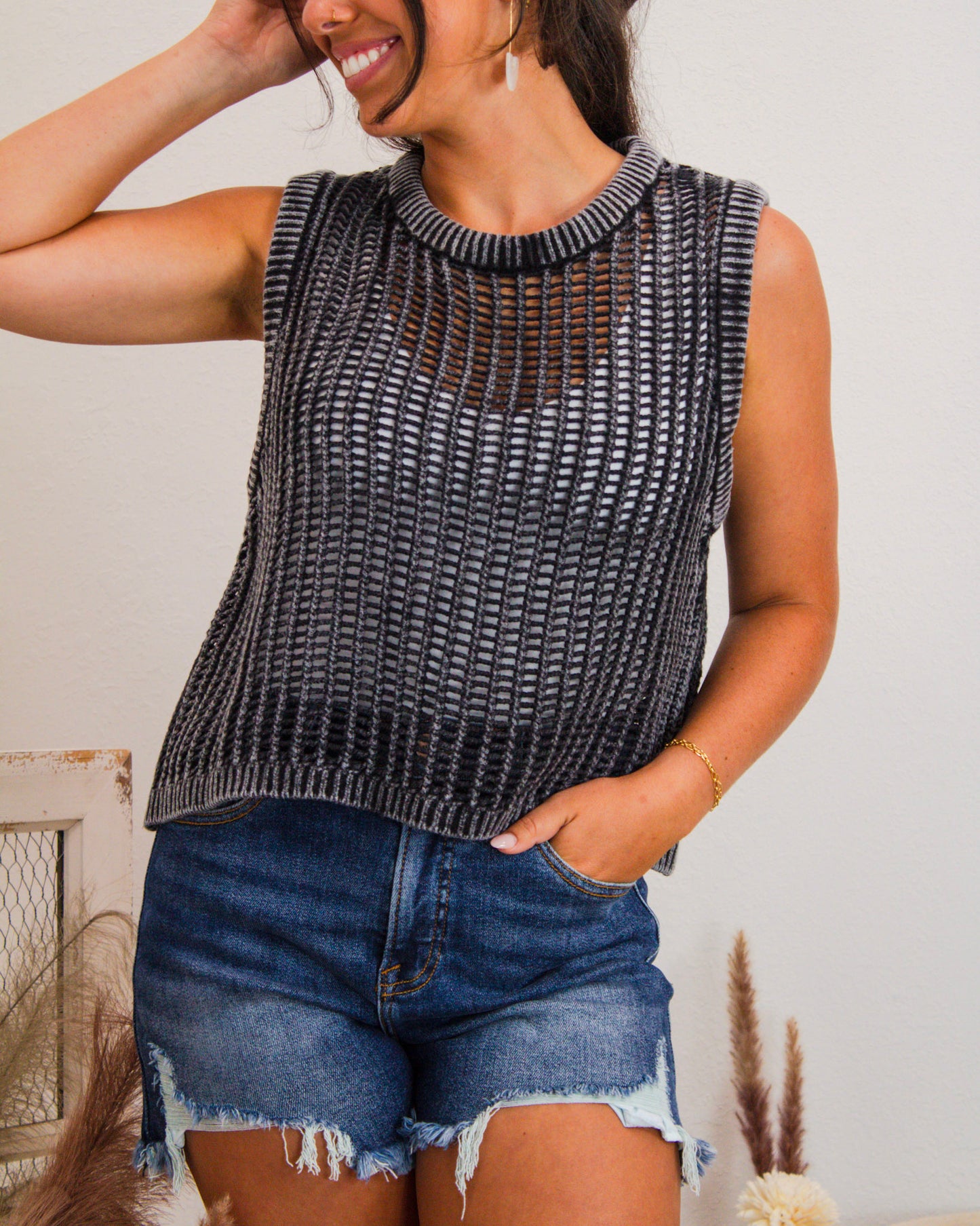Woman wearing a black open-knit sleeveless sweater tank layered over a black bralette, paired with high-rise distressed denim shorts for a trendy summer look.