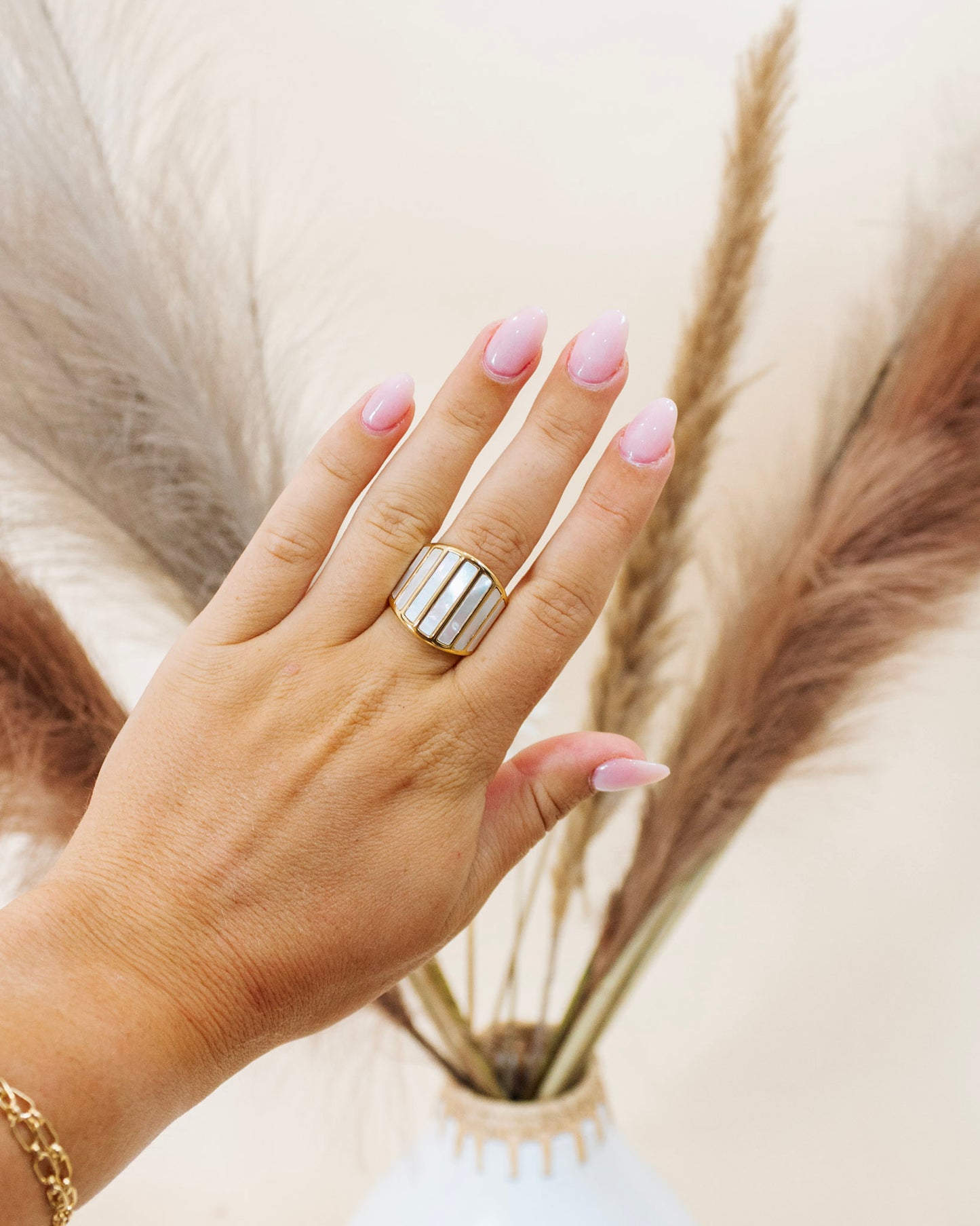 Hand wearing a bold gold statement ring with vertical mother-of-pearl inlays, styled against pampas grass on a neutral background