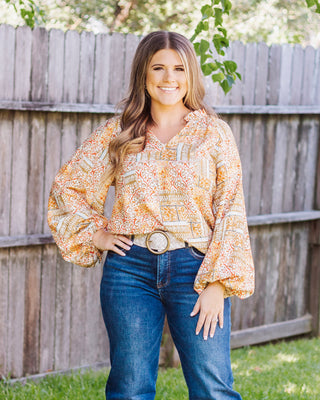 A full-body shot of a woman smiling in a colorful patterned blouse and blue straight-leg jeans against a wooden fence.