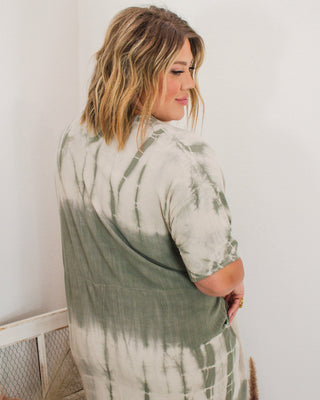 Woman wearing a sage green and cream tie-dye midi dress with short sleeves, viewed from the back, styled in a boutique setting.