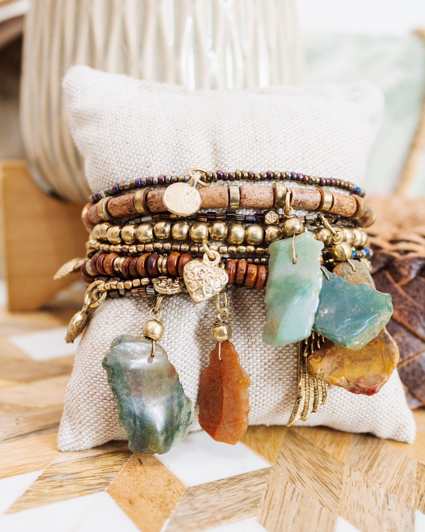 Stack of boho gemstone charm bracelets with mixed beads, wood accents, and gold detailing displayed on a neutral cushion.