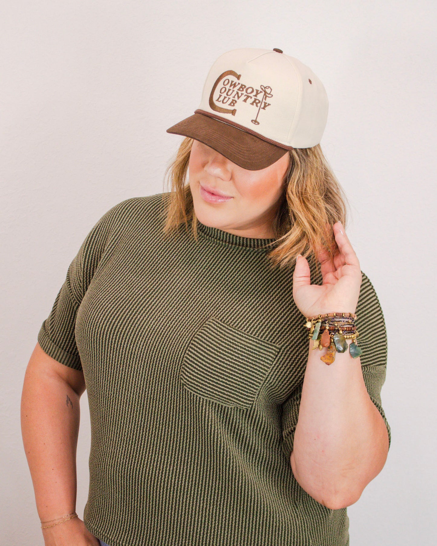 Woman wearing a “Cowboy Country Club” trucker hat, olive ribbed top with chest pocket, and stacked gemstone charm bracelets.