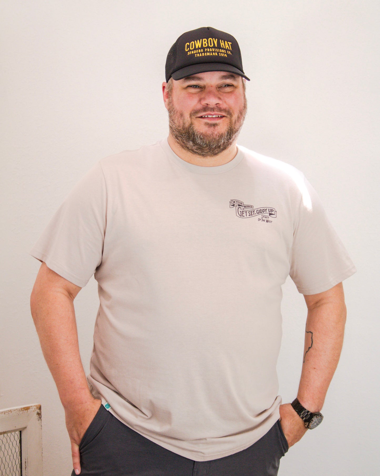 Man wearing a light beige Sendero Provisions Co. t-shirt with “Get Set, Giddy Up” text on the chest, paired with a black “Cowboy Hat” trucker cap and dark shorts.