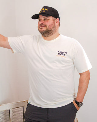 Man wearing a cream Sendero Provisions Co. t-shirt with “Daggum Mirage” text on the chest, styled with a black trucker hat and dark shorts for a casual look.
