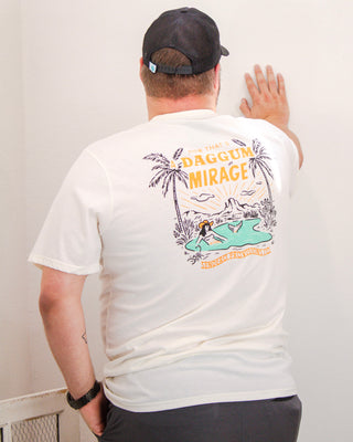 Man wearing a cream Sendero Provisions Co. t-shirt with a colorful back graphic of a desert oasis, palm trees, and “Daggum Mirage” text, styled with a black cap.