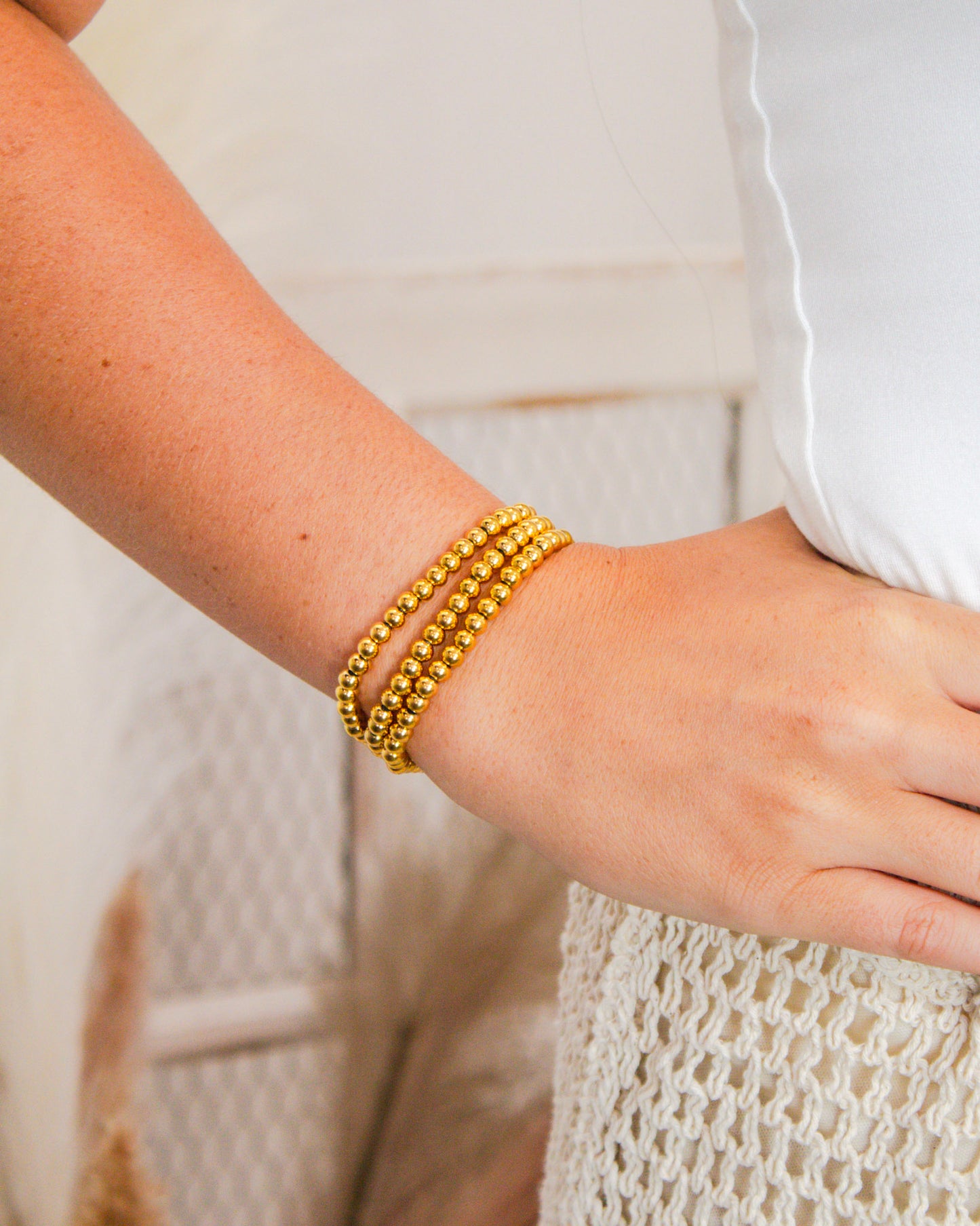 Close-up of gold beaded stretch bracelets stacked on a woman's wrist, paired with a white textured outfit.