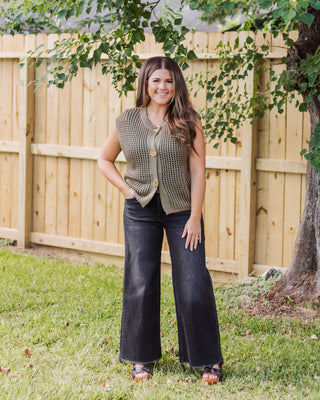 Woman wearing an olive green sleeveless knit vest with gold statement buttons, paired with high-waisted black wide-leg jeans and platform sandals.