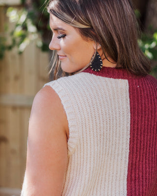 Close-up of a woman wearing a sleeveless ribbed knit colorblock sweater vest in cream and rust tones, paired with black teardrop leather earrings.