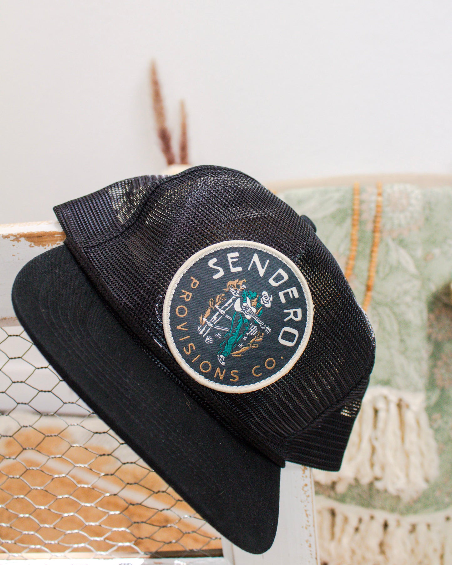 Black mesh trucker hat from Sendero Provisions Co. with a round patch featuring a cowboy illustration, displayed on a rustic backdrop with neutral decor.