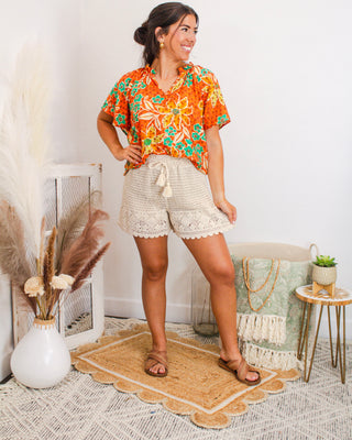 Woman wearing a vibrant orange floral button-up blouse with teal and yellow accents, paired with cream crochet scalloped shorts and tan sandals for a breezy summer look.