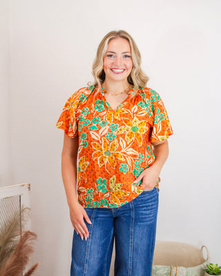Woman wearing a bright orange short-sleeve floral blouse with yellow and teal flower patterns, styled with blue denim jeans for a cheerful, casual summer look.